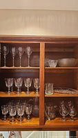 Wide view of wooden cabinet shelf with various glassware including wine glasses, bowls, plates, and pitchers arranged inside.