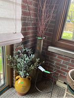 Wide view showing both ceramic jugs with faux plants and metal plant stand near brick wall and window.