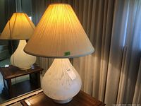 Full view of the lamp on a wooden table with a mirror behind, showing the beige pleated cloth shade and the frosted glass base with speckled pattern.