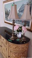 Side view of both matching bronze candle lamps with fabric shades and a glass vase with artificial pink and white flowers on a wooden cabinet.