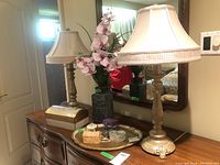 Photo showing two metal table lamps with white cloth shades on a wooden dresser with a mirror, a floral arrangement with pink orchids, a jewelry box, and a tray with several small trinket boxes.