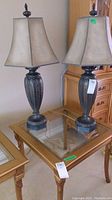 Two matching ornate base table lamps with beige fabric shades on a glass-top wooden side table.