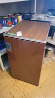 Full view of the brown Danby vintage mini fridge with wood grain top panel, showing front and top sides, placed on a floor with cans and kitchen items behind it.