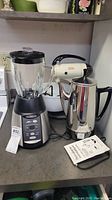 Sunbeam mixer, Oster blender, and stainless steel percolator arranged on kitchen countertop alongside product tags and manual