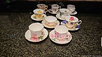 Photo showing nine assorted teacups and saucers with various floral designs, mostly with matching saucers, arranged on a dark countertop.