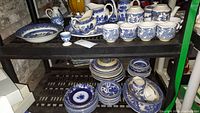 Wide angle view of two shelves holding multiple Blue Willow pattern dinnerware pieces including plates, cups, and serving dishes.