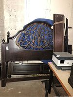 Disassembled king bed frame including a decorative headboard with metal insert, footboard, and sideboards leaning against the wall in a warehouse setting.