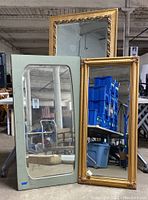 Three large gold framed wall mirrors displayed leaning against warehouse furniture and storage. One rectangular mirror has curved edges in frame design, others are straight rectangular with floral/leaf patterned carved frames.
