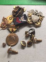 Overall view showing a bronze medal with standing figure, several pairs of cufflinks, a ring, a small round pin, and a blue cloth military rank insignia patch with eagle and chevron.