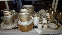 Full collection shot of Noritake Goldridge china including stacked cups, saucers, plates, serving dishes, teapot, creamer, and sugar bowl, all with white base and gold detailing.