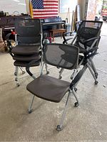Front and angled view of multiple office chairs stacked and arranged in warehouse setting on concrete floor. Chairs have black mesh backs, black seats, and silver metal frames with wheels and armrests.