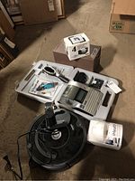 View of all items in the lot including Brother P-Touch label maker in case, iRobot Roomba vacuum, iHealth Blood Pressure Dock in box, and other items on floor.