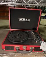 Front view of the Victrola portable record player showing the open lid with Victrola logo, platter, tonearm, control knobs, and built-in speaker grille on a red suitcase-style case.