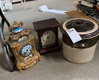 Photo showing two vintage mantel clocks and a 2-gallon farmhouse crock with lid on a concrete floor.