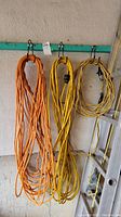 Three coiled extension cords hanging on hooks against a textured wall, showing orange, yellow, and yellow-black stripe cords with plugs visibly attached.