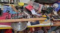 Wide view of workbench with mixed hardware items including circular saw blades, hand tools, nuts, bolts, and packaged hydraulic jack.