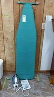 Full view of teal fabric covered folding ironing board standing upright, iron, and instruction manual on floor in front of wood panel wall.