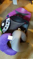 Photo showing two travel neck pillows (tan and purple) and two Dr. Ho's Perfectback Rests (black and magenta) with massage ball features and mesh backing placed on carpet.