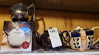 Three teapots on a wooden shelf: stainless steel Paderno teapot on its box, dark brown ceramic spherical teapot, hexagonal ceramic teapot with blue and orange floral pattern.