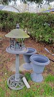 Photo shows two large dark gray plastic planter urns fluted with pedestal bases and a tall metal bird feeder with ornamental base and roofed hanging cage feeder.