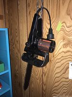 Black Remington electric chainsaw hanging on chain against wooden panel wall, side view showing motor and handle with attached power cord