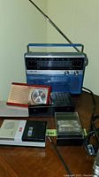 Blue and silver Candle solid state radio with telescoping antenna, along with a red and beige Philips transistor radio with analog clock, black Philips cassette recorder, and other small items on wooden surface.