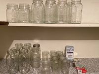 Photo showing multiple clear glass canning jars of different sizes arranged on shelves and a counter, some with lids and some without.