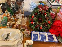 Wide shot of Christmas-themed items: miniature Christmas trees, figurines, decorative wreath, teacups on platter, packaged items, and a red sack.