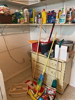 Wide shot of cleaning supplies on shelves with mop handles leaning against a chest and litter box on floor