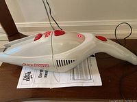 White Dirt Devil cordless handheld vacuum with red buttons and logo, shown on floor with power cord plugged in and a red indicator light illuminated.