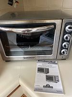 Front view of stainless steel KitchenAid countertop oven with glass door and chrome handle. Four control knobs visible on right side and original instruction manual placed underneath.