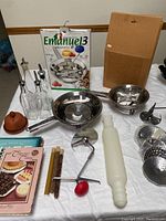 Photo of whole lot showing two stainless steel food mills, Emanuel 3 box, extra blades, glass oil and vinegar dispensers, cookbooks and various kitchen tools on tabletop.