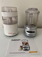 Photo showing both Cuisinart food processor and Sunbeam citrus juicer along with the food processor's instruction manual on white surface.