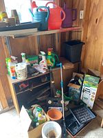 Wide view of garden supplies including watering cans, sprayers, plant pots, fertilizers, and small gardening tools in a wooden shed corner.