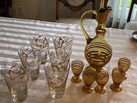 Photo showing six clear glasses decorated with gold rose bouquet design and six smaller amber striped liqueur glasses alongside a large amber and clear striped pitcher with handle.