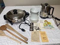 Photo showing the stainless steel automatic skillet with electric cord, three wooden utensils, three white bowls, stainless steel drip coffee maker, and various informational booklets and instructions spread on a white tablecloth.