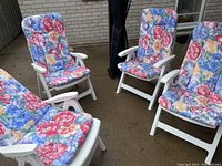 Four white resin reclining chairs arranged together on a patio with floral cushions attached.