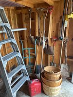 Full view showing the stepladder, long-handled shovels, spade, rake, rototiller attachment, dolly, gas can, and wooden bushel baskets inside a wooden shed