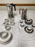 Photo showing the large and smaller stovetop espresso makers alongside 11 cups and saucers on a white cloth surface.