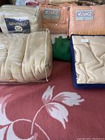Photo showing five wool comforters and bed covers in beige and pink, including Eaton and Royal Touch brands, with two in plastic packaging.