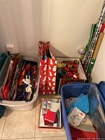 Full view of plastic bins containing assorted gift bags, tissue paper, ribbons, and bows with rolls of wrapping paper on the side