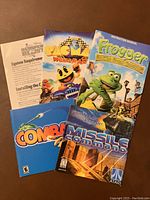 Top-down photo of six game instruction manuals and cover pages showing titles such as Bubble Burst, Pac Man World Rally, Frogger The Great Quest, Combat, Missile Command.