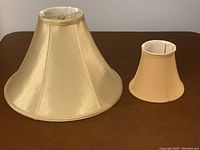 Two fabric lamp shades, one large cream color and one smaller beige, standing next to each other