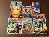 Front view of seven paperback comic books showing full cover art with characters including Iron Man, X-Men teams, Captain America, and Robotech.