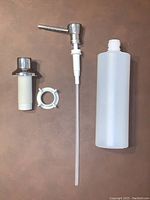 Disassembled soap dispenser components including chrome pump, white mounting collar, mounting nut, and semi-translucent refill bottle with long dip tube.