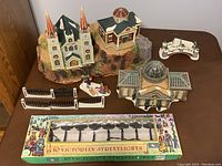 Full overview of Winter Village 12-piece figurine set displayed on table with church, city hall, Margrove building with glued trees, fence sections, bridge with lamp, and children tobogganing scene
