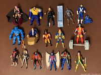 Front view of the 19 X-Men action figures arranged on a flat surface showing characters including Psylocke, Strong Guy, Long Shot, Iceman, Cyclops, Beast, Professor X, Wolverine, Banshee, Colossus, Nightcrawler, Storm, Gambit, Archangel, Bishop, and Rogue along with several accessories such as a mechanical arm, chair, and base stand.
