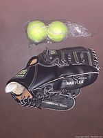 Black Mizuno youth baseball glove labeled Max Flex Professional Model, includes two yellow tennis balls wrapped in plastic on a brown surface.