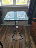 Front angle view of one square glass top patio table with metal tripod base showing overall structure and condition needing cleaning.