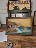 Framed ocean landscape painting showing sea, rocks, and sunset with a yellow sky and blue water.